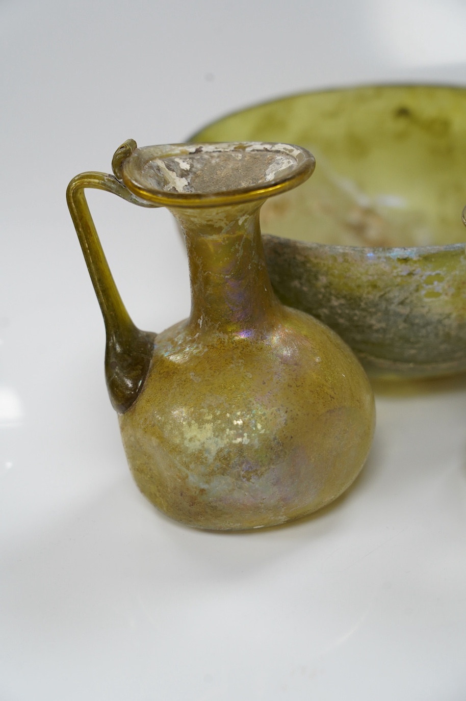 Three items of Roman glassware to include a bowl, a miniature jug and a vessel, bowl 14cm diameter. Condition - bowl repaired